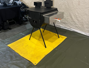 A yellow fireproof mat placed under a black stove, situated on a gray floor inside a tent environment.
